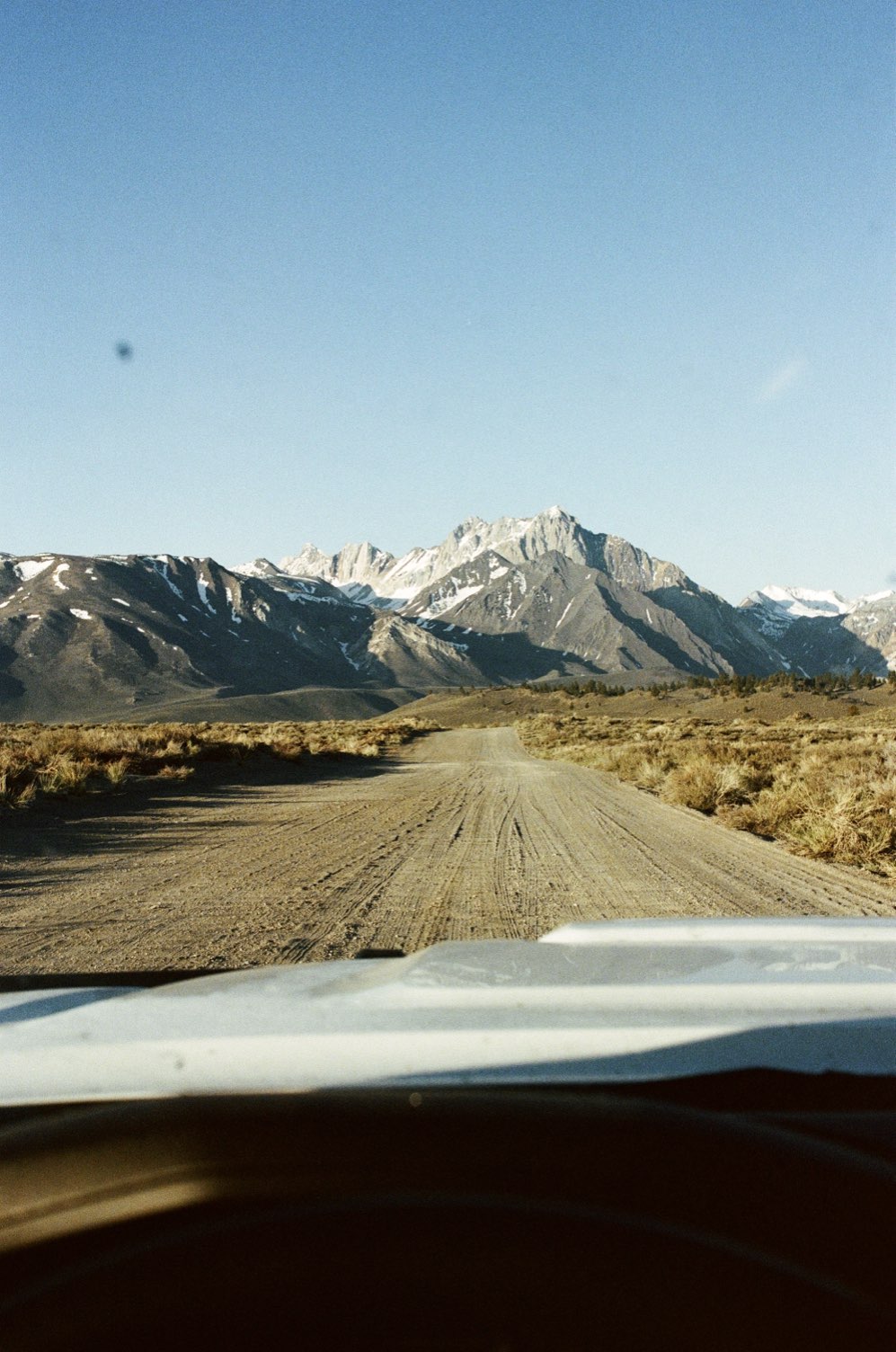 A dirt road leading toward snowy peaks, seen over a car hood