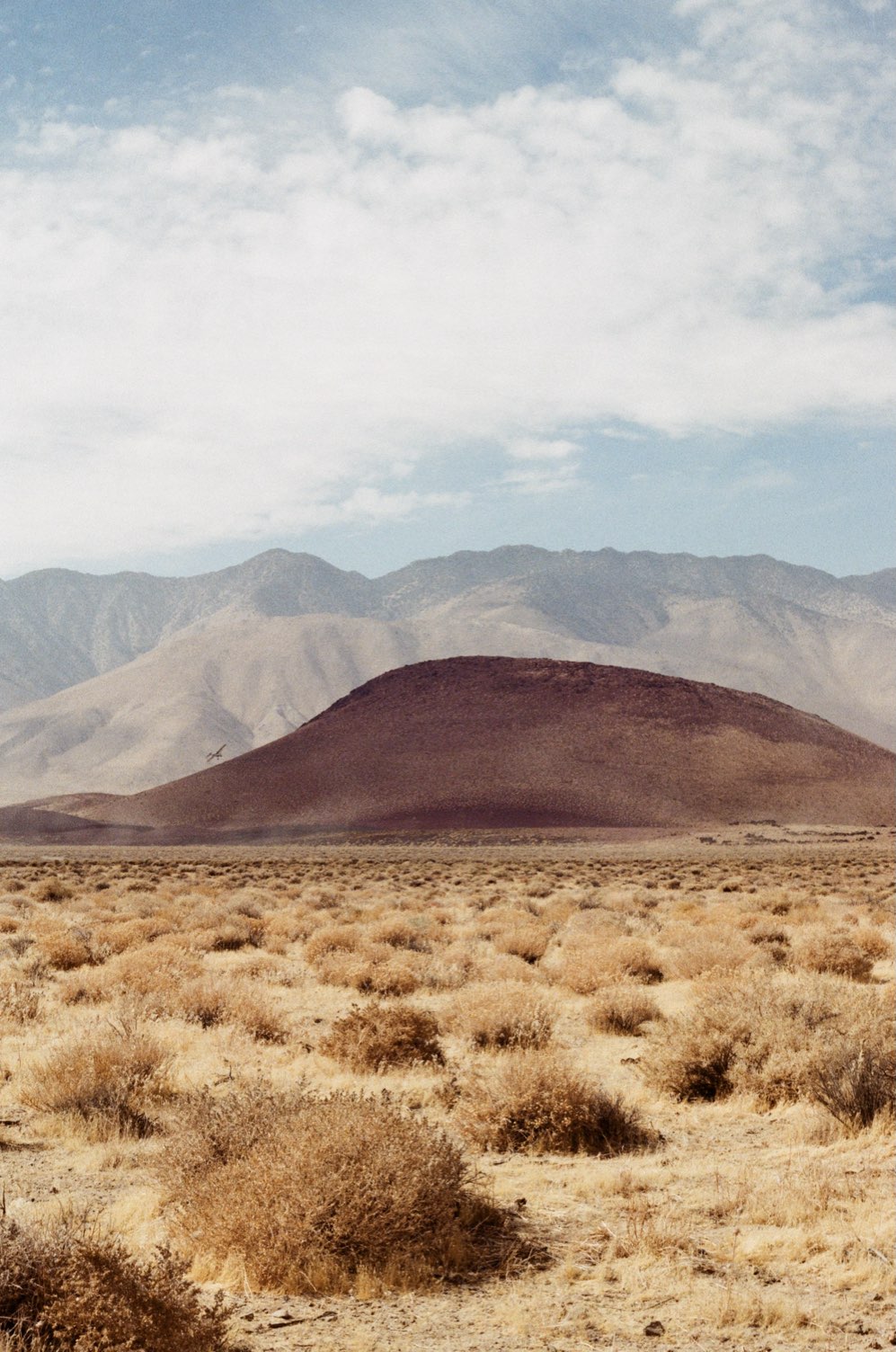A red cinder cone in high desert scrub below pale mountains